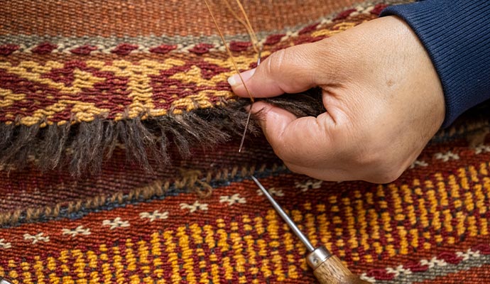 A close-up of a specialist's hands using a needle and thread to perform detailed fringe repair on a patterned oriental rug
